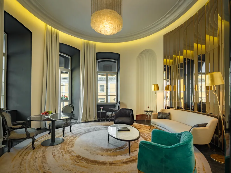 Elegancki salon hotelowy w hotelu De Paris z okrągłym układem przestrzeni. Pomieszczenie wyposażone jest w kremową sofę, szmaragdowy fotel aksamitny oraz czarny fotel wypoczynkowy ustawione wokół owalnego stolika kawowego z marmurowym blatem. Po lewej stronie znajduje się okrągły stół z czarnym szklanym blatem z owocami i stylowe krzesła w stylu barokowym. Wnętrze zdobią wysokie okna z kremowymi zasłonami, złote lampy podłogowe, mozaikowa ściana z luster w złotej oprawie oraz kryształowy żyrandol. Podłogę pokrywa okrągły dywan w beżowo-złotych odcieniach. Sufit z ozdobnym gzymsem podświetlony jest ciepłym, żółtym światłem ambientowym.